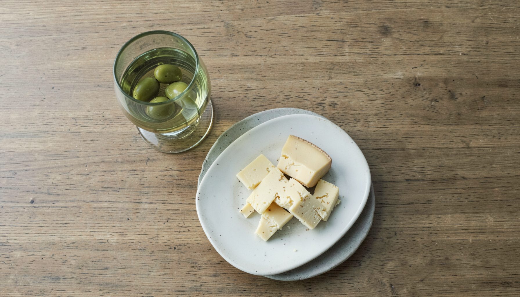 Overhead flat-lay photograph of a wooden table with a glass of vinho verde, a plate of cheese, and a small bowl of olives, soft natural light, muted greens and browns.